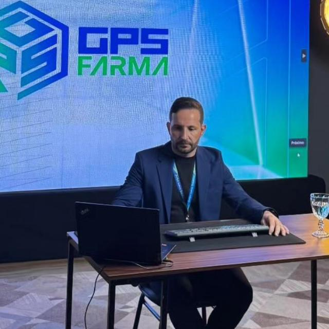 Foto do Marcos sentado a uma mesa com notebook, em frente a um telão com a marca GPS Farma.